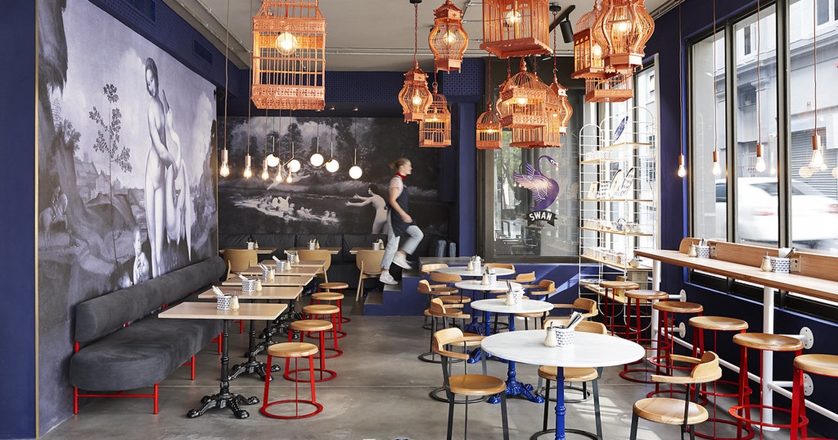Interior de cafetería con mesas, ventanales y lámparas, en plano abierto
